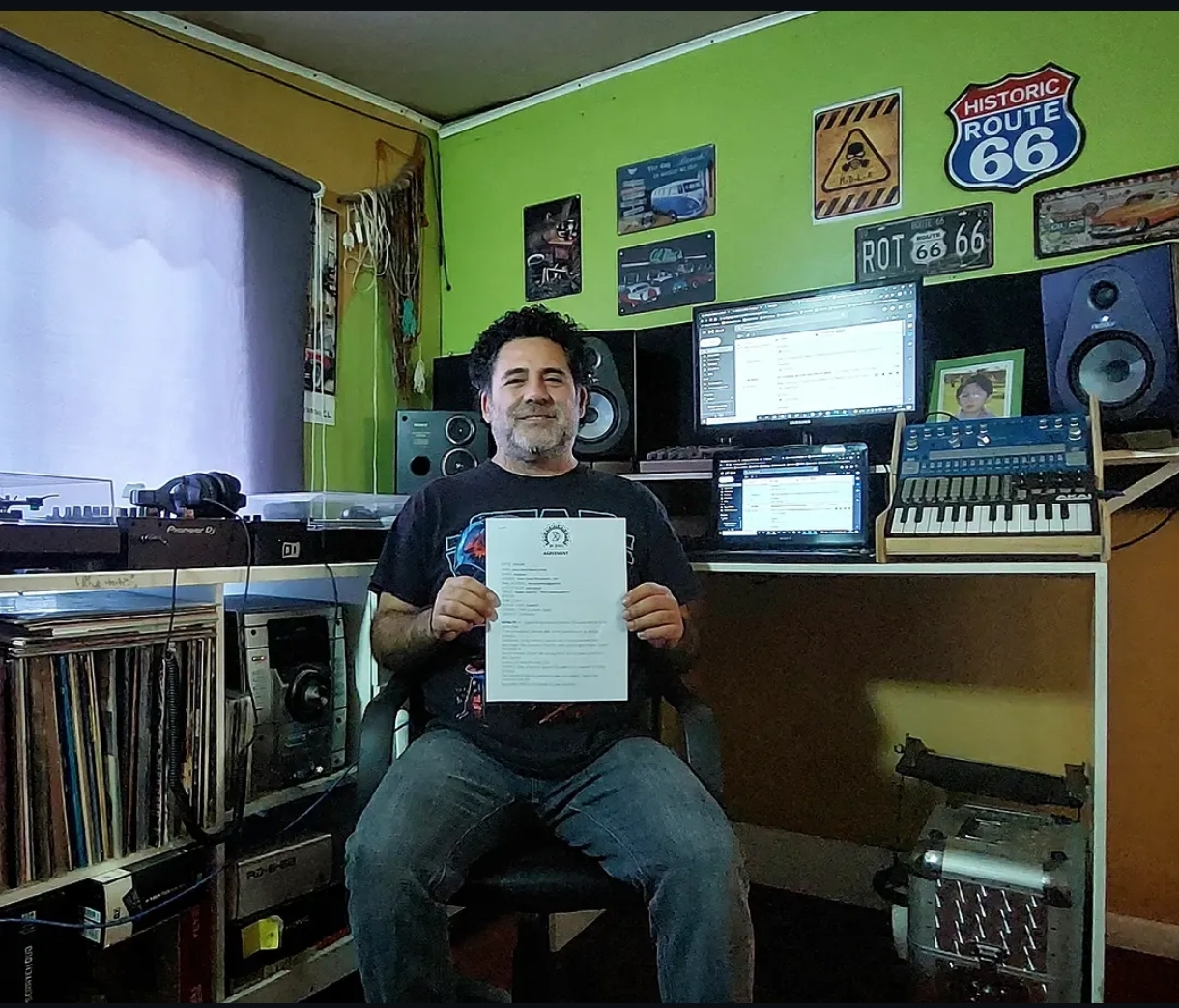 Henry Riquelme sentado en su estudio, sosteniendo un documento frente a sus equipos musicales.