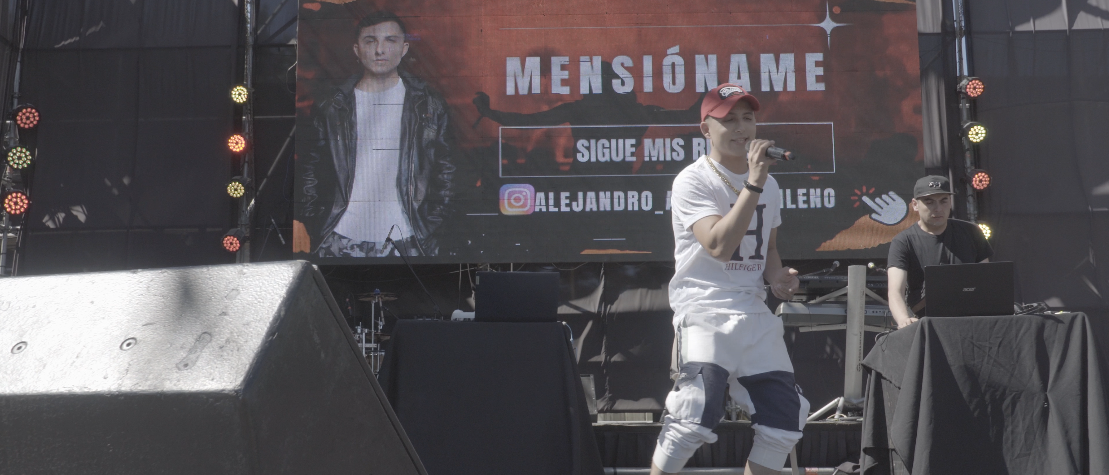 Alejandro Araya canta con gorra roja y micrófono frente a una pantalla con su imagen y sus redes sociales.