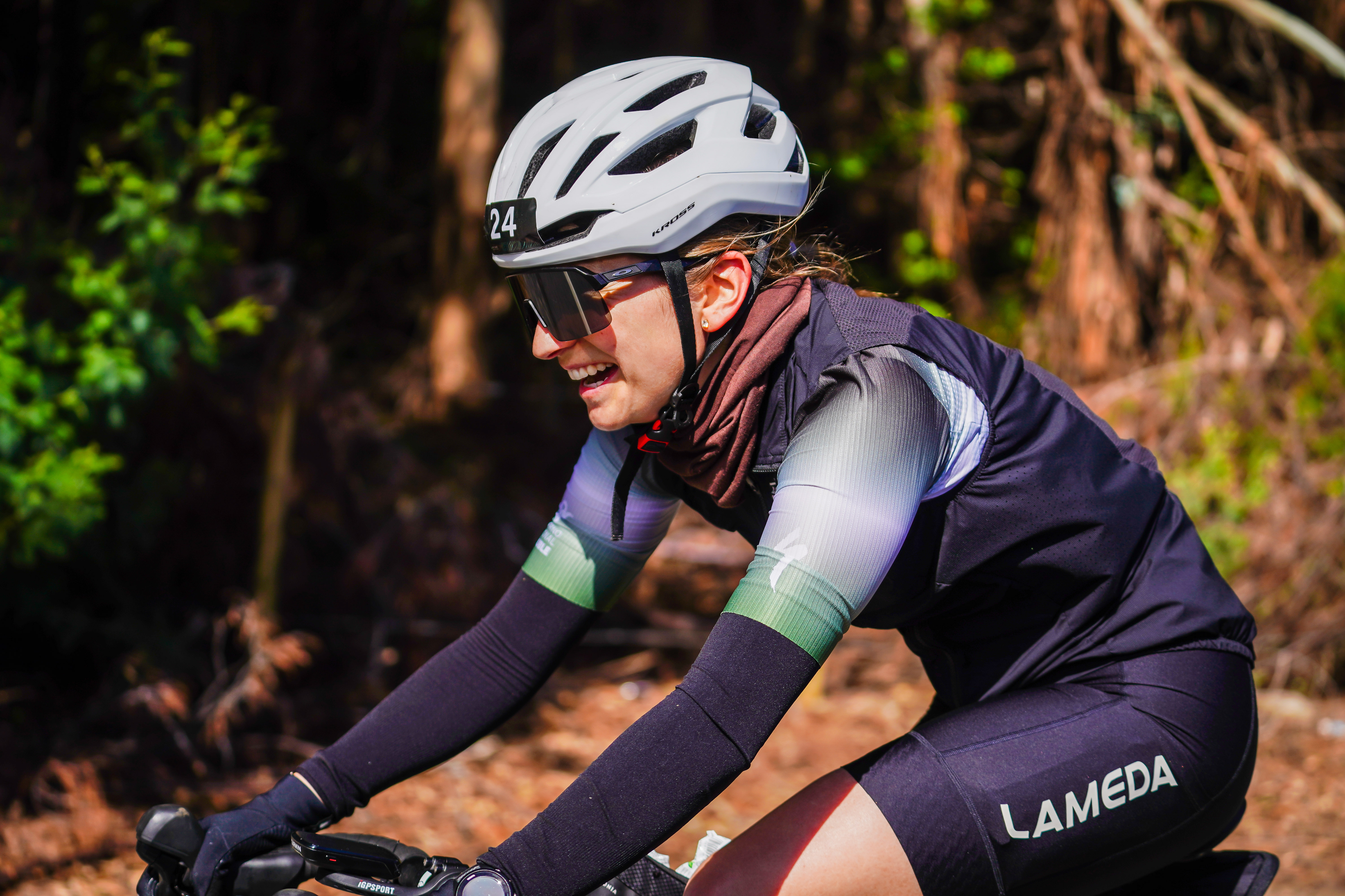 Una ciclista con casco blanco, gafas y camiseta del Gran Fondo pedalea junto a una zona arbolada.