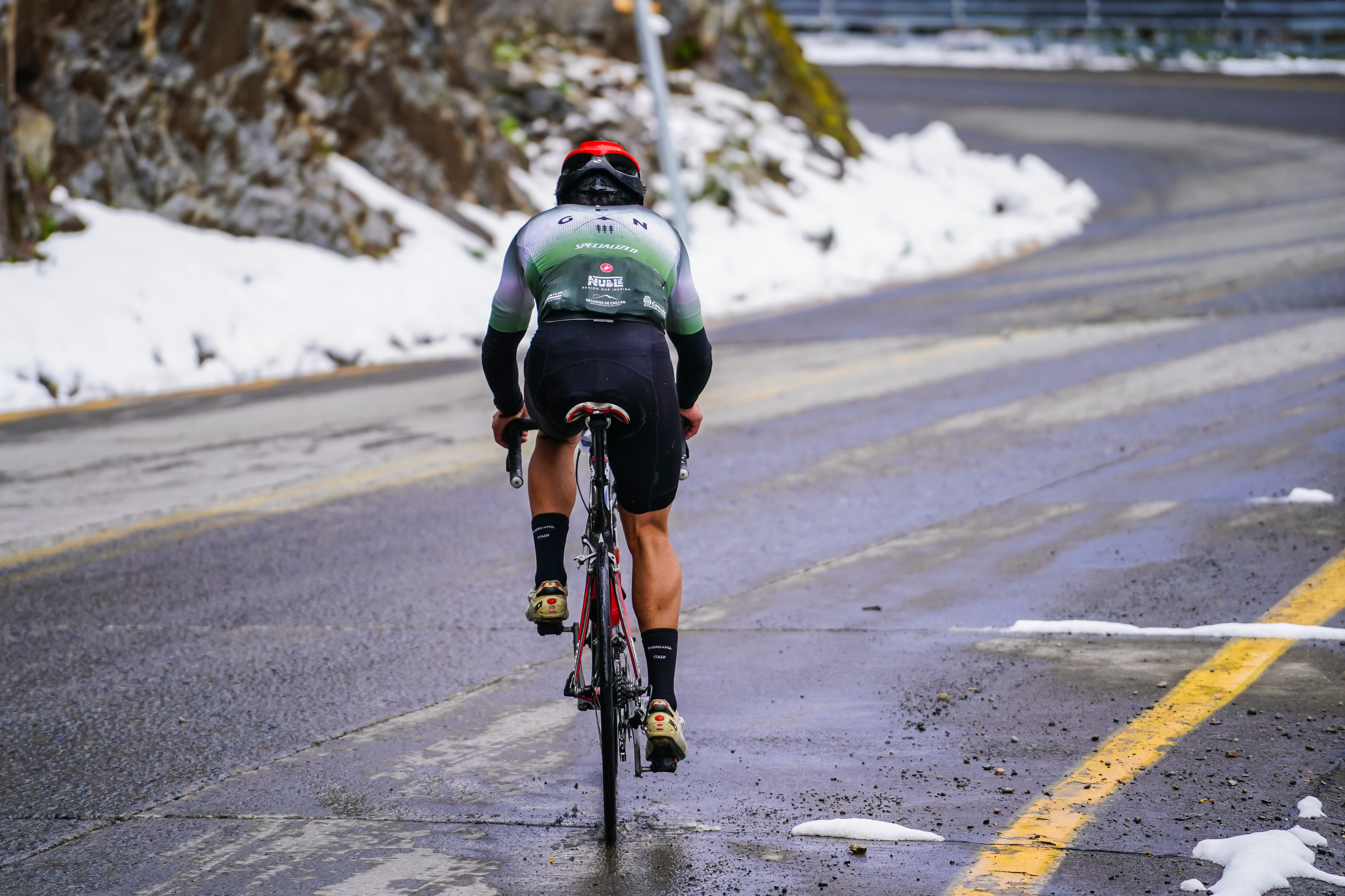 Ciclista de espaldas en una carretera de montaña que curva hacia la derecha, con nieve a ambos costados.